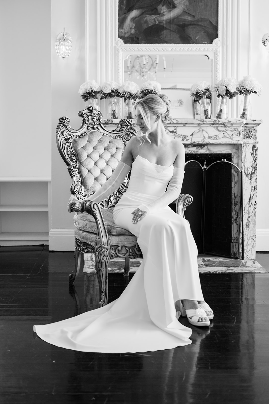 Black and white portrait of the bride sitting elegantly in a vintage chair beside a fireplace mantel.
